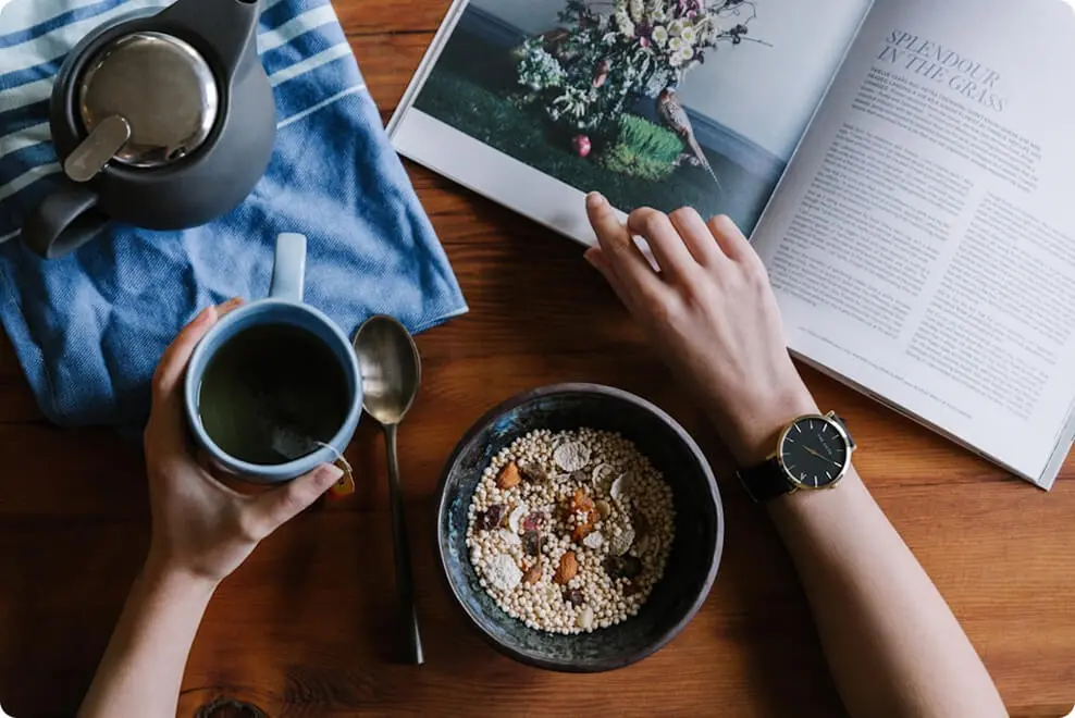 Foto como foco nas mãos femininas em cima de uma mesa, fazendo uma leitura e tomando café com granola